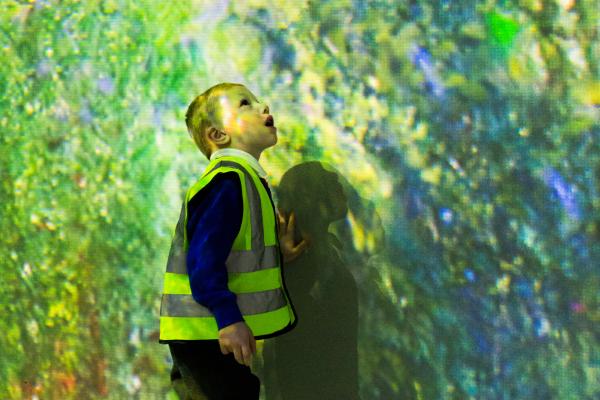 A young child wearing a high-visibility vest gazes in awe at the immersive artwork surrounding him in the FRAMELESS gallery.