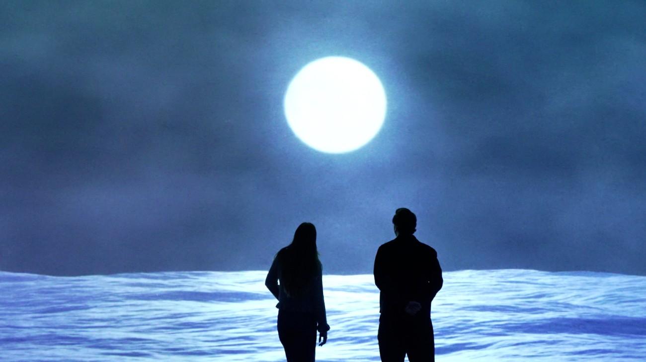 A man and a woman standing in front of a projected image of a full moon above the sea