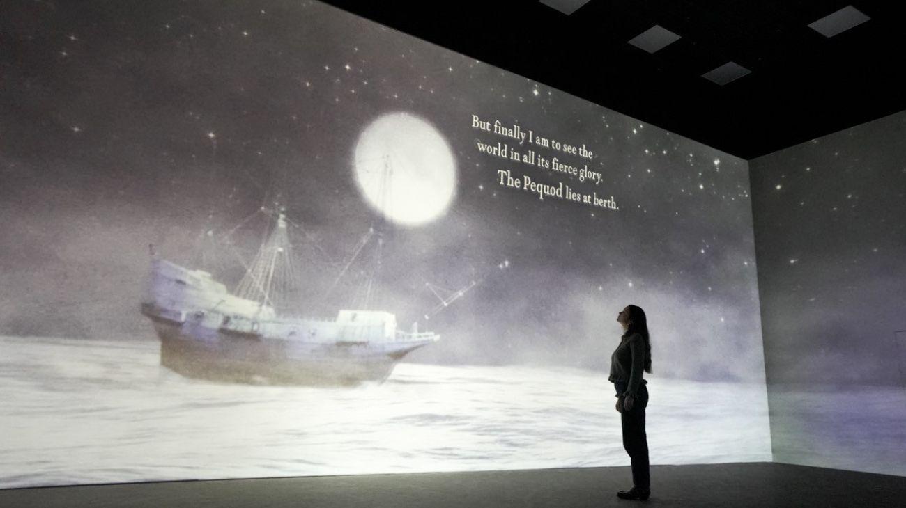 A woman standing in front of a projected image of a ship at sea with a full moon above it and words on the screen.