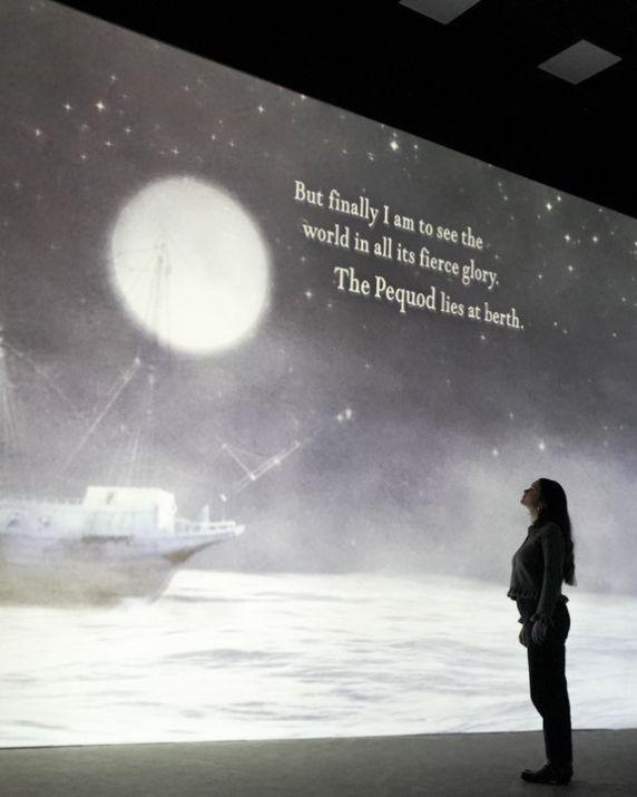 A woman standing in front of a projected image of a ship at sea with a full moon above it and words on the screen.