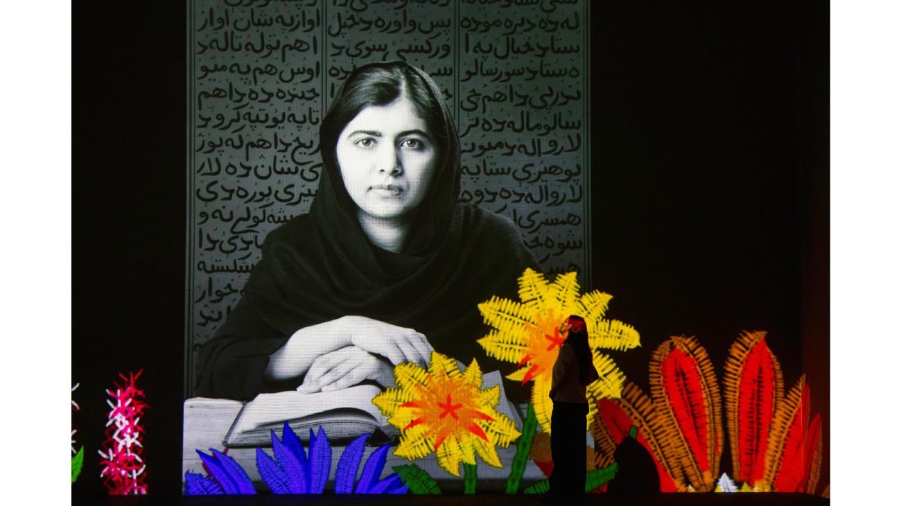 The silhouette of a woman standing in front of a portrait of Malala Yousafzai with colourful flowers also showing on the Gallery wall.