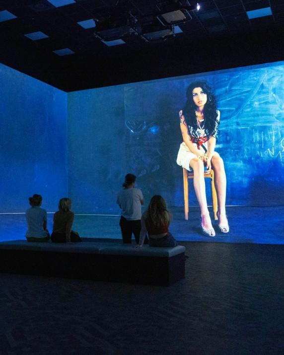 A group of people in front of a portrait of Amy Winehouse sat on a chair in front of a blue background.