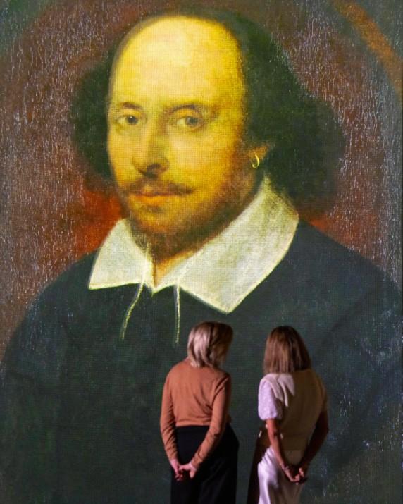 Two women standing in front of a portrait of William Shakespeare