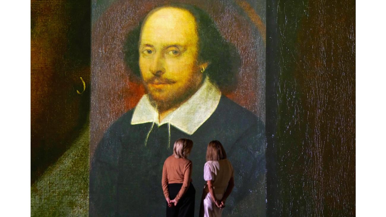 Two women standing in front of a portrait of William Shakespeare